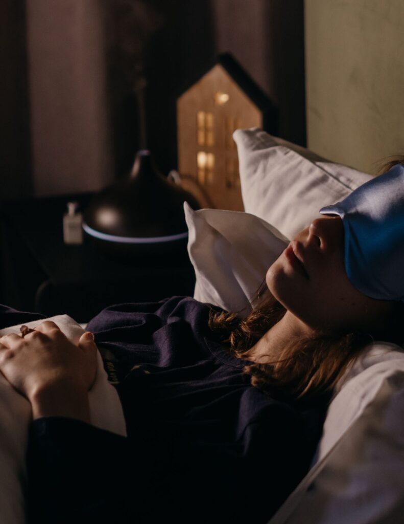 Eine Person liegt im Bett mit einer Schlafmaske und einem Kissen, umgeben von Bettwäsche.